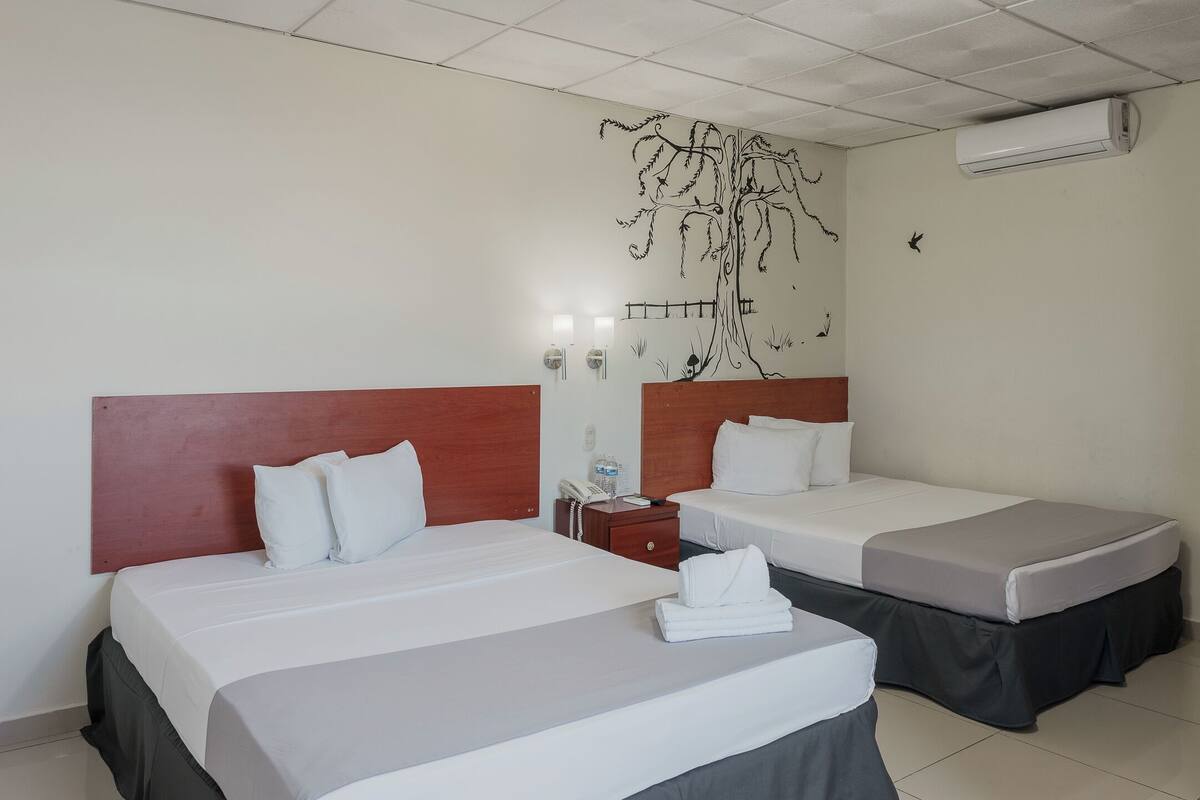 Habitación de hotel con dos camas, cabecero de madera, un mural en la pared, mesita de noche y unidad de aire acondicionado.