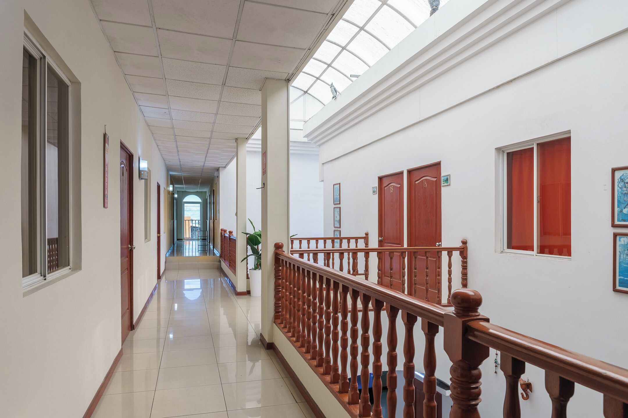 A long, narrow hallway with white walls and wooden doors, leading to a bright room with a large window.