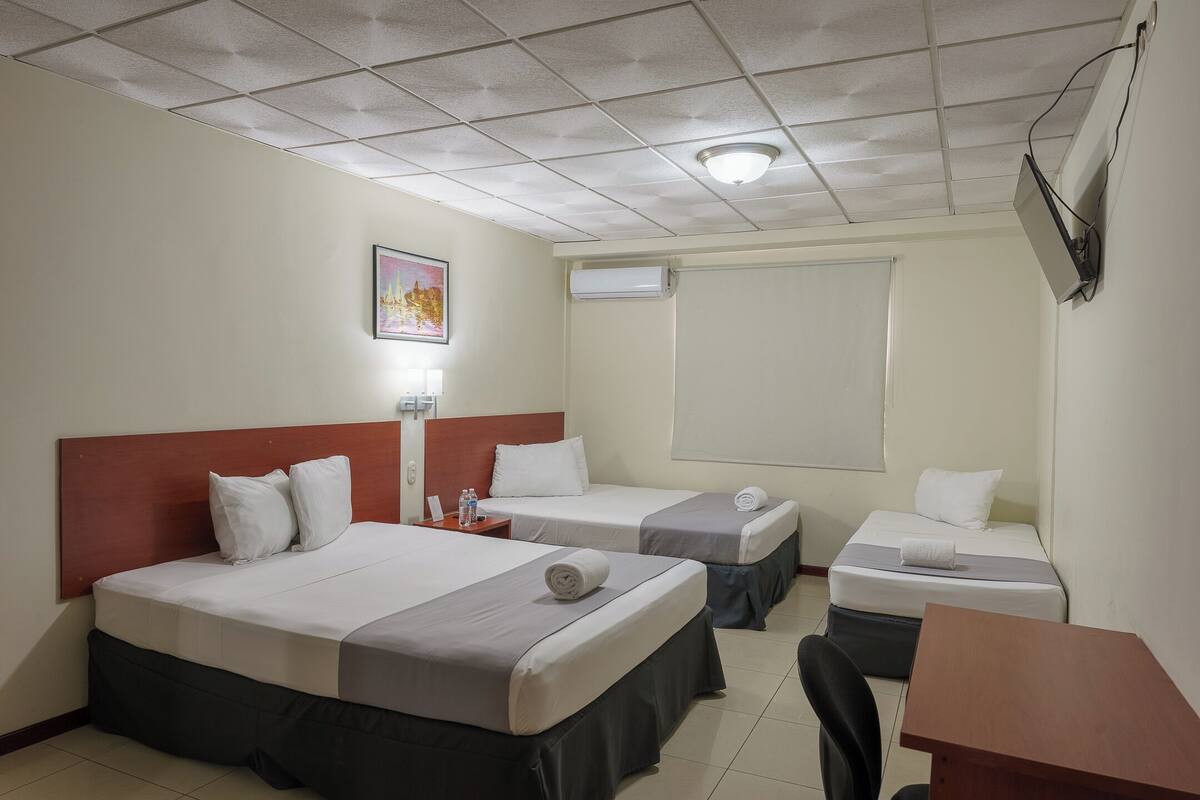 Habitación de hotel con dos camas, un escritorio, una silla, una mesita de noche, una lámpara y un equipo de aire acondicionado.