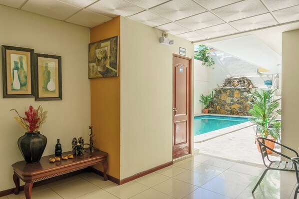 A hotel lobby with a reception desk, framed artwork, a door labeled "BAÑO," and a view of an outdoor swimming pool area.