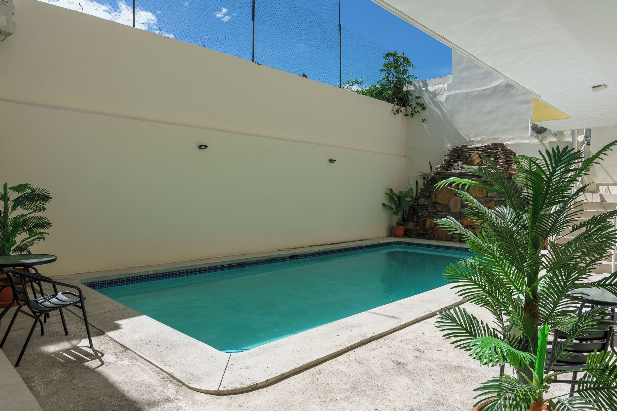 Una piscina rectangular amb un videt de rajola, envoltada per un sòl de formigó. Hi ha dues taules amb cadires, una a cada banda de la piscina. Un aulet blanc ofereix ombra sobre part de la zona de la piscina.