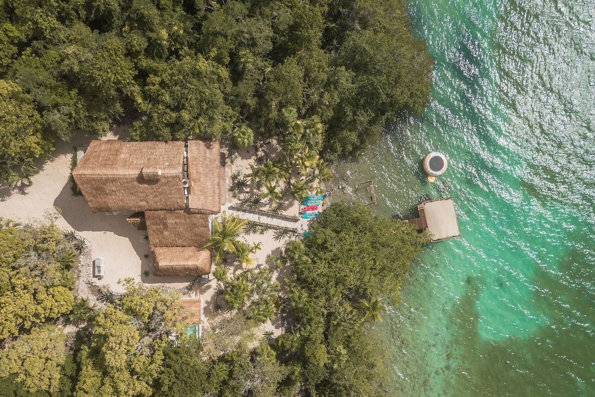 Luftaufnahme eines Anwesens am Strand mit strohgedeckten Gebäuden, einem Schwimmbereich mit Booten und einem klaren, türkisblauen Meer.