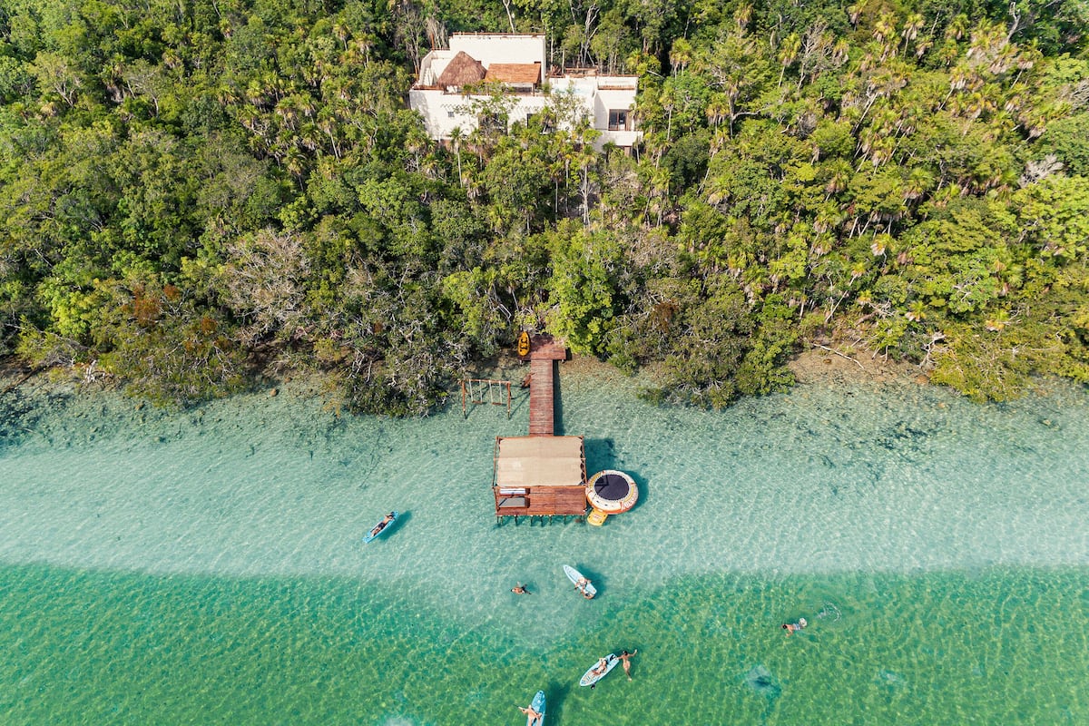Ein Luftbild einer Strandlandschaft mit einer Holzsteganlage, klarem türkisblauem Wasser und einem kleinen Haus im Hintergrund.