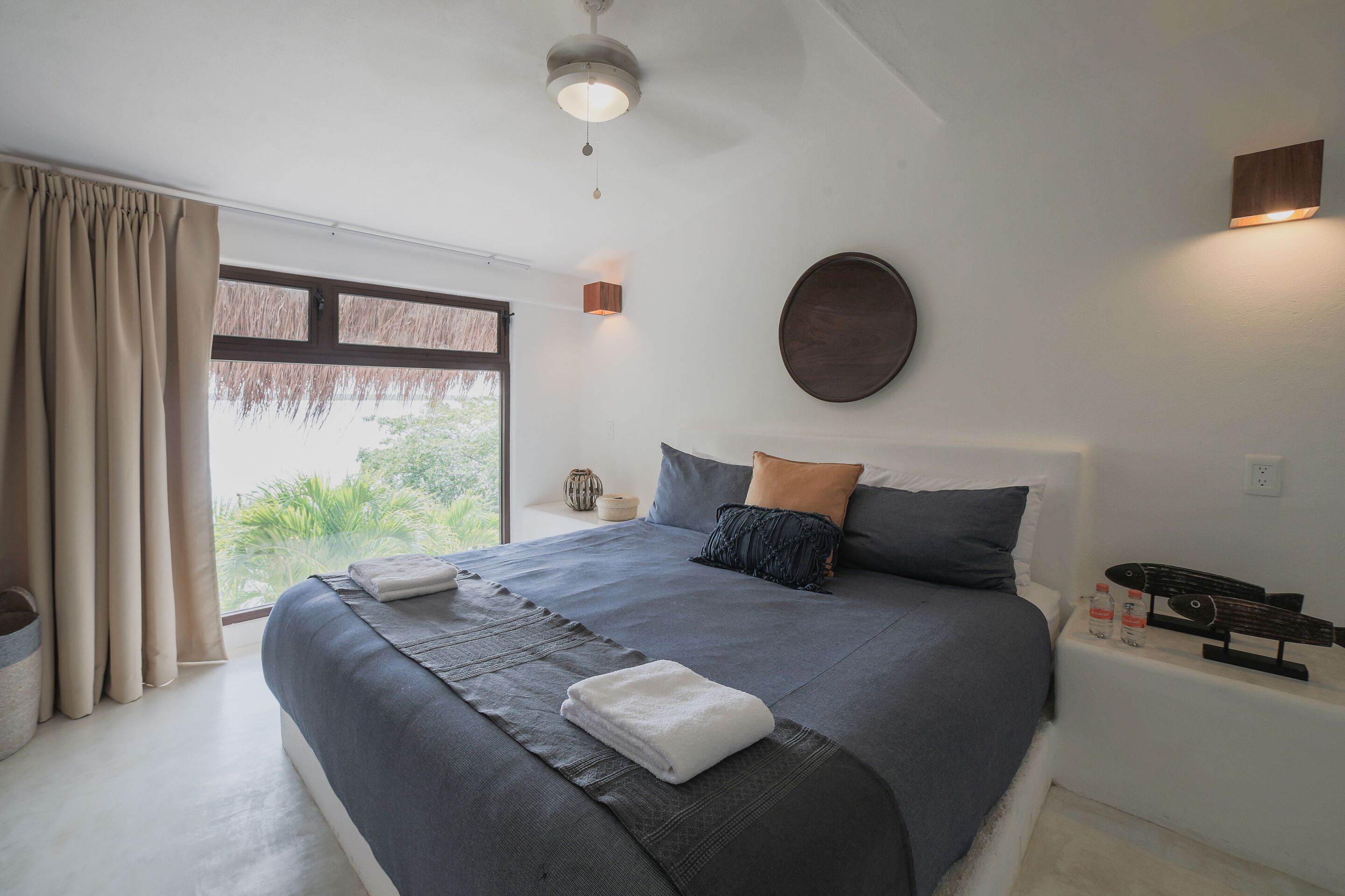 Una cama bien hecha con cubrecama gris y azul, una manta blanca y una almohada. Junto a la cama hay una mesita con una botella de agua y un monopatín.