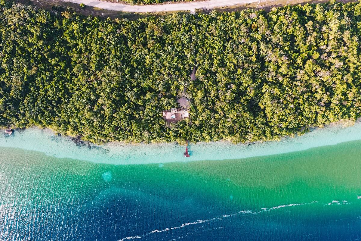 Eine Vogelperspektive zeigt ein Küstenanwesen mit dichtem Wald, einem klaren Strand und einem kleinen Haus am Wasserrand.