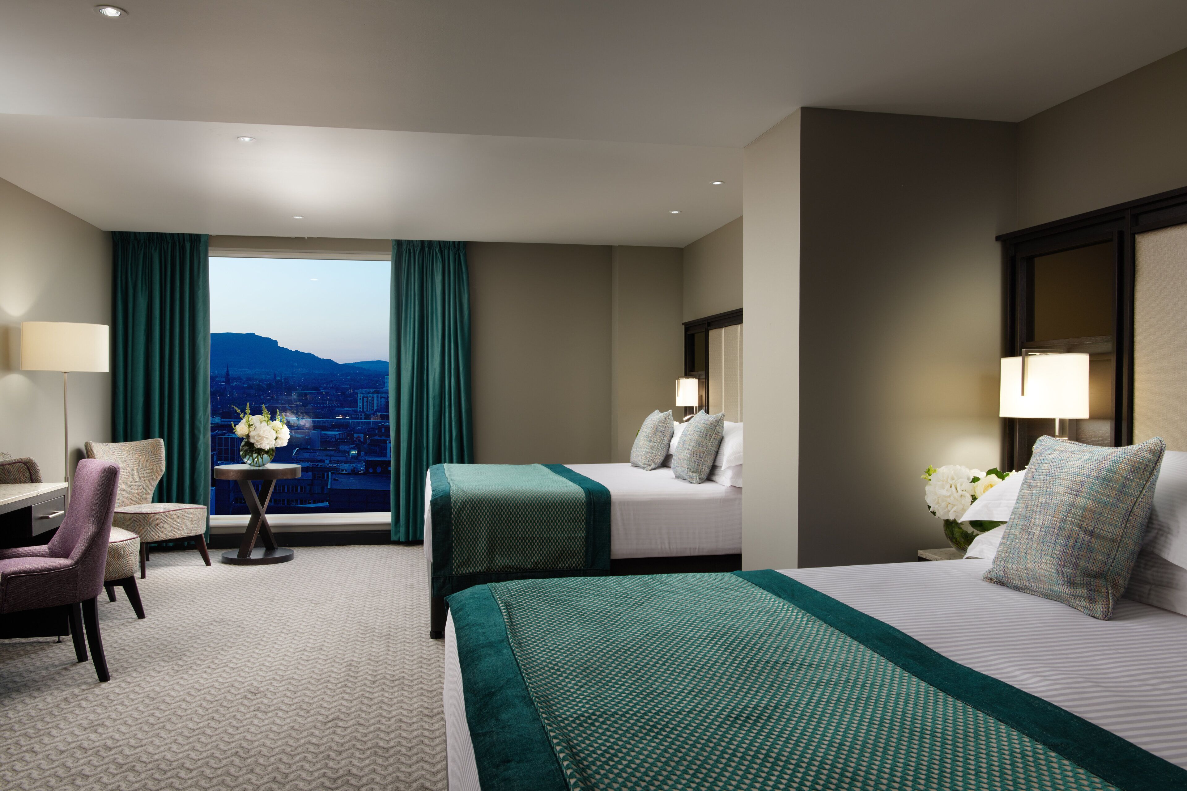 A hotel room with a bed, a desk, a chair, a window with curtains, and a view of a cityscape.
