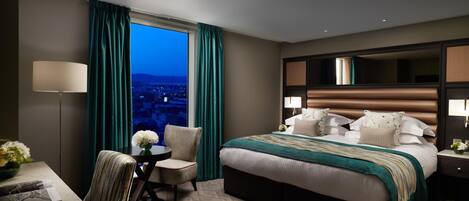 A hotel room with a large bed, a desk with a chair, a small table with a vase of flowers, and a view of the city at dusk.