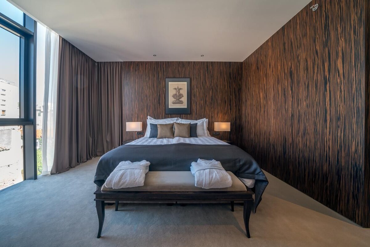 A modern hotel room with a large bed, two bedside lamps, a framed artwork above the bed, and a view through a large window.