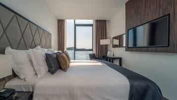 Et hotelværelse med en stor seng, et fladskærms-tv monteret på væggen, et skrivebord med en lampe, og udsigt til vandet udenfor.