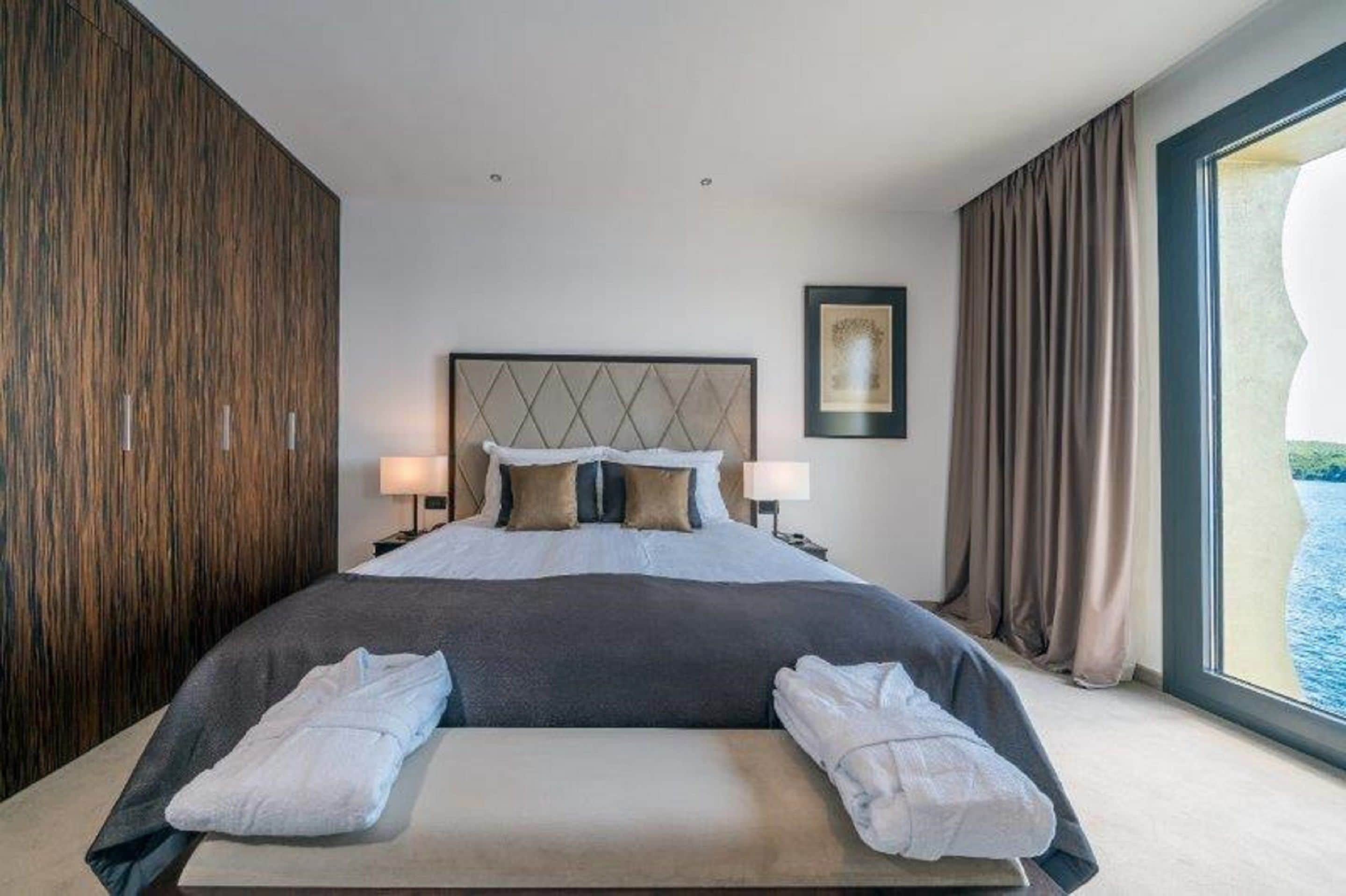 Ein modernes Hotelzimmer mit einem großen Bett, zwei Nachttischlampen, einem gerahmten Bild an der Wand und Meerblick durch ein großes Fenster.