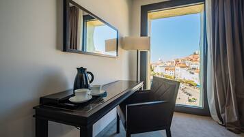 Habitación de hotel con escritorio, una silla, cafetera y vistas a un paisaje urbano a través de un ventanal grande.