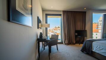 A hotel room with a bed, a desk with a chair, a TV, and a large window with a view of buildings.