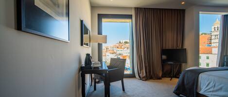 A hotel room with a bed, a desk with a chair, a TV, and a large window with a view of buildings.