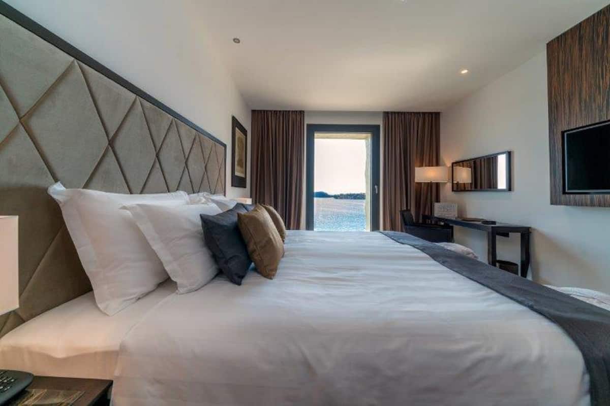 Une chambre d’hôtel avec un grand lit, un bureau, une télévision et une vue sur la mer, visible par la porte.