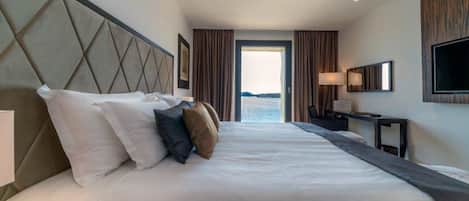 Une chambre d’hôtel avec un grand lit, un bureau, une télévision et une vue sur la mer, visible par la porte.