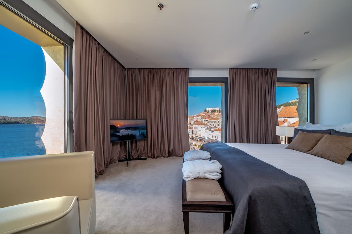 Une chambre d’hôtel avec un grand lit, un banc, une télévision et une vue sur la mer et le paysage urbain.