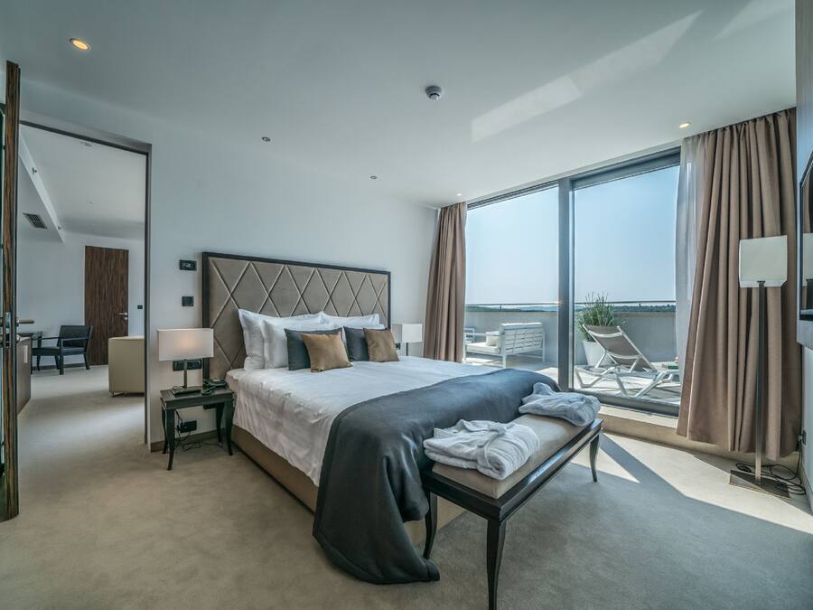 A modern hotel room with a large bed, a bedside table with a lamp, a view of the balcony, and a chair.