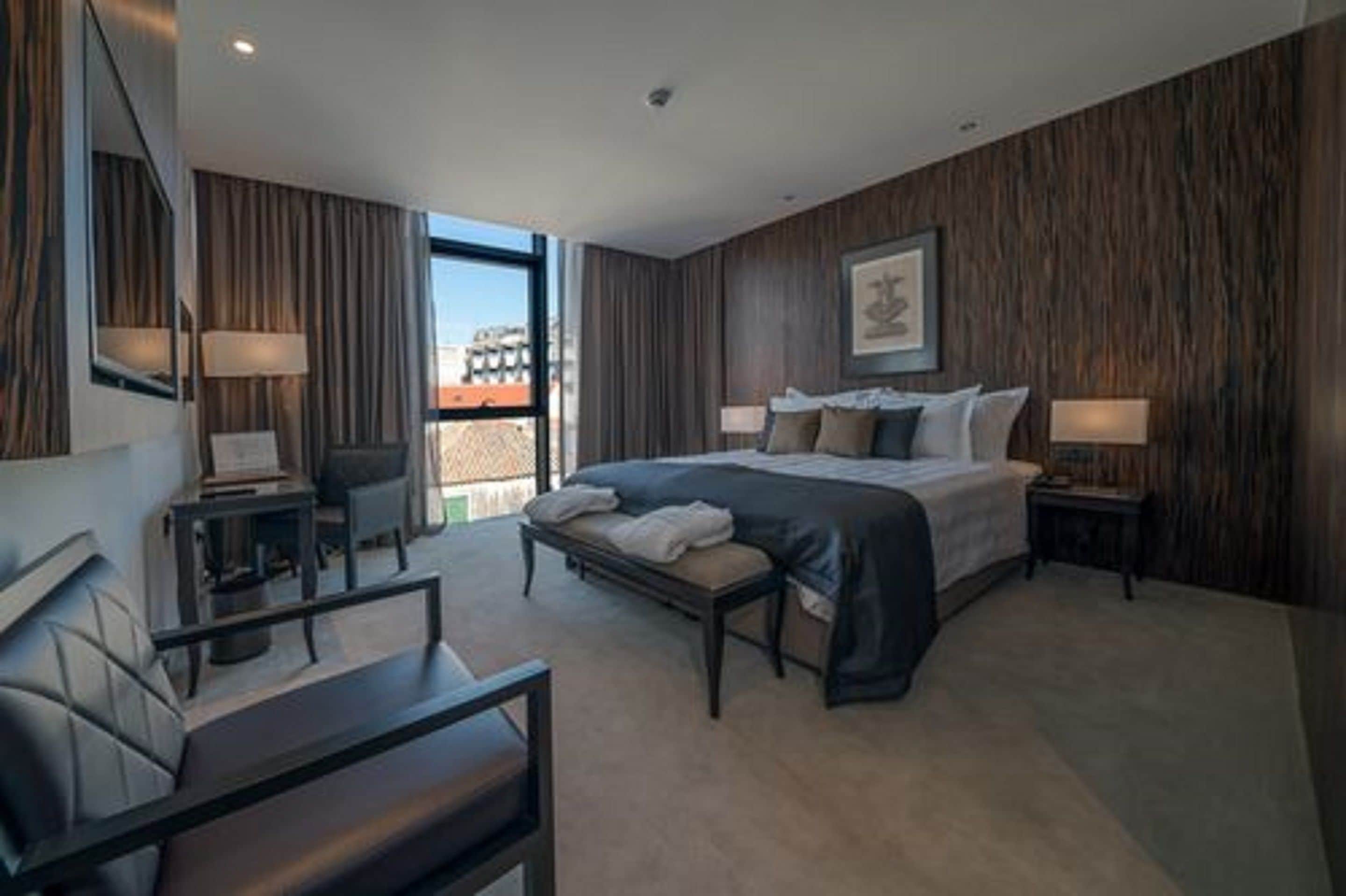 A modern hotel room with a large bed, a desk, and a chair. There is a window with a view of buildings outside.