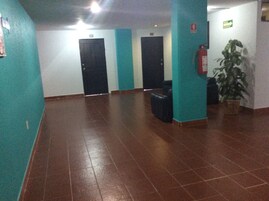 Un pasillo con paredes verde azulado, piso de baldosas marrones y puertas negras. A la derecha, hay una planta en maceta y un extintor.
