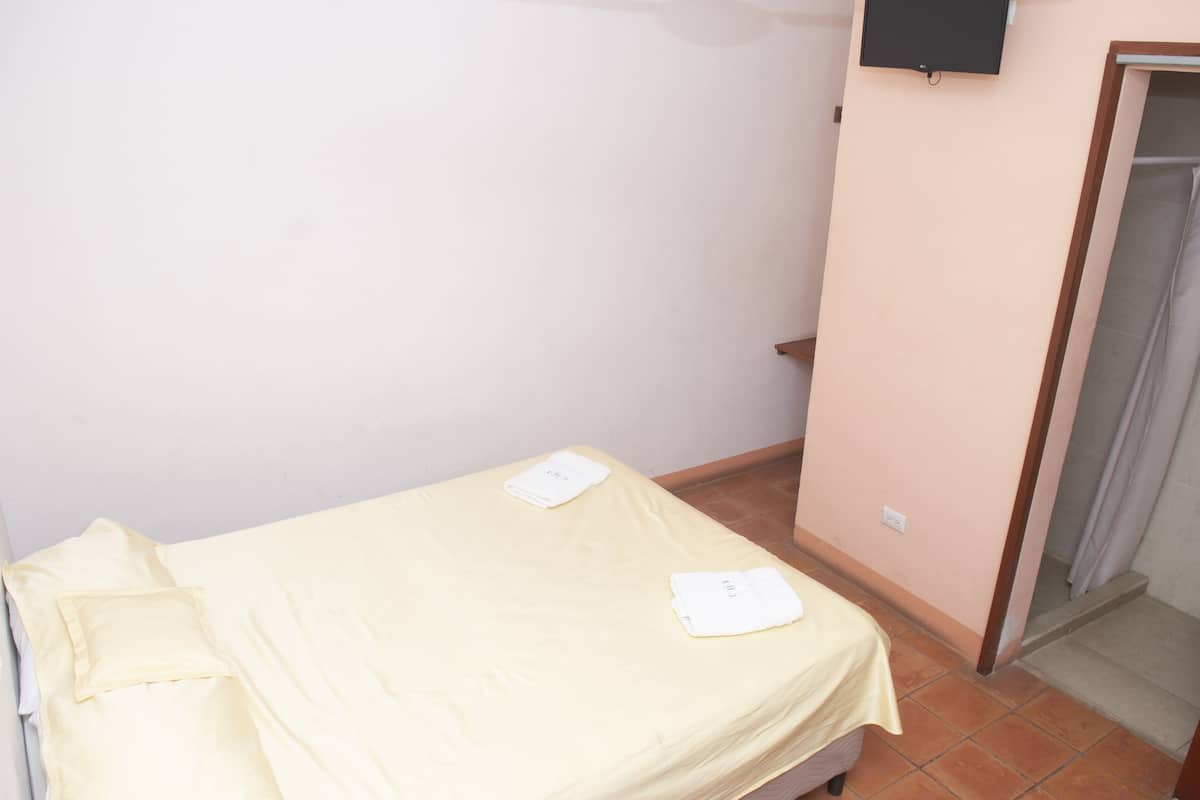 A small, neatly made bed with a plain yellow bedspread and two pillows, a wall-mounted TV, and a door leading to a bathroom.
