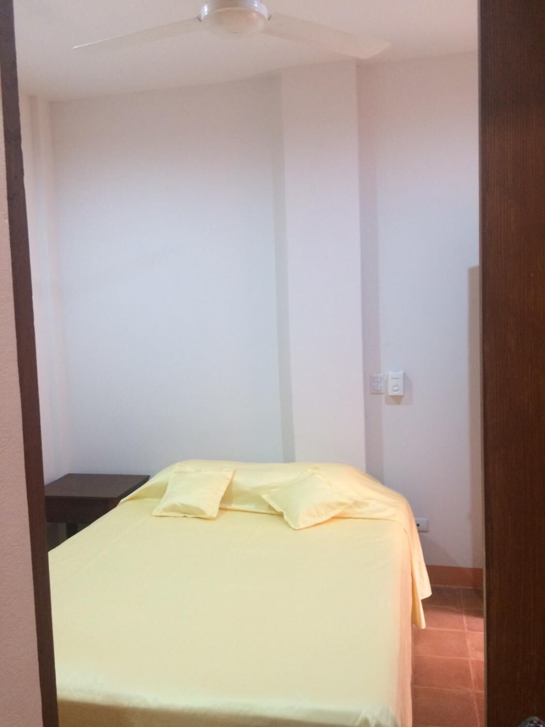 Una habitación pequeña con una cama individual, pared blanca lisa y ventilador de techo.