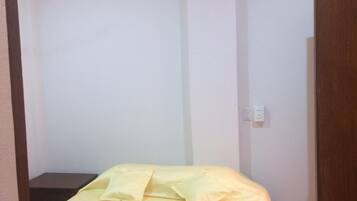 Una habitación pequeña con una cama individual, pared blanca lisa y ventilador de techo.