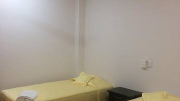 Quarto de hotel com duas camas de solteiro, cada uma com colcha amarela e toalha branca. Há um ventilador de teto sobre as camas, e as paredes são de cor branca lisa.