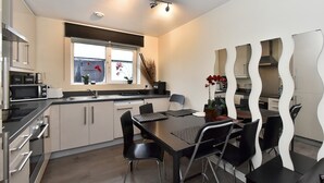 A modern kitchen with a dining area, featuring stainless steel appliances, a window with flowers, and a unique wavy mirror design. - Belgravia Apartments- Wilton Road (London)