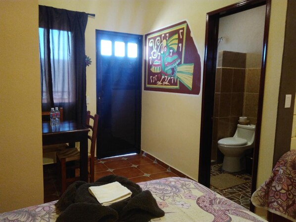 Une petite chambre d’hôtel comprenant un lit, un bureau, une chaise, une salle de bain et une porte ornée d’une affiche.