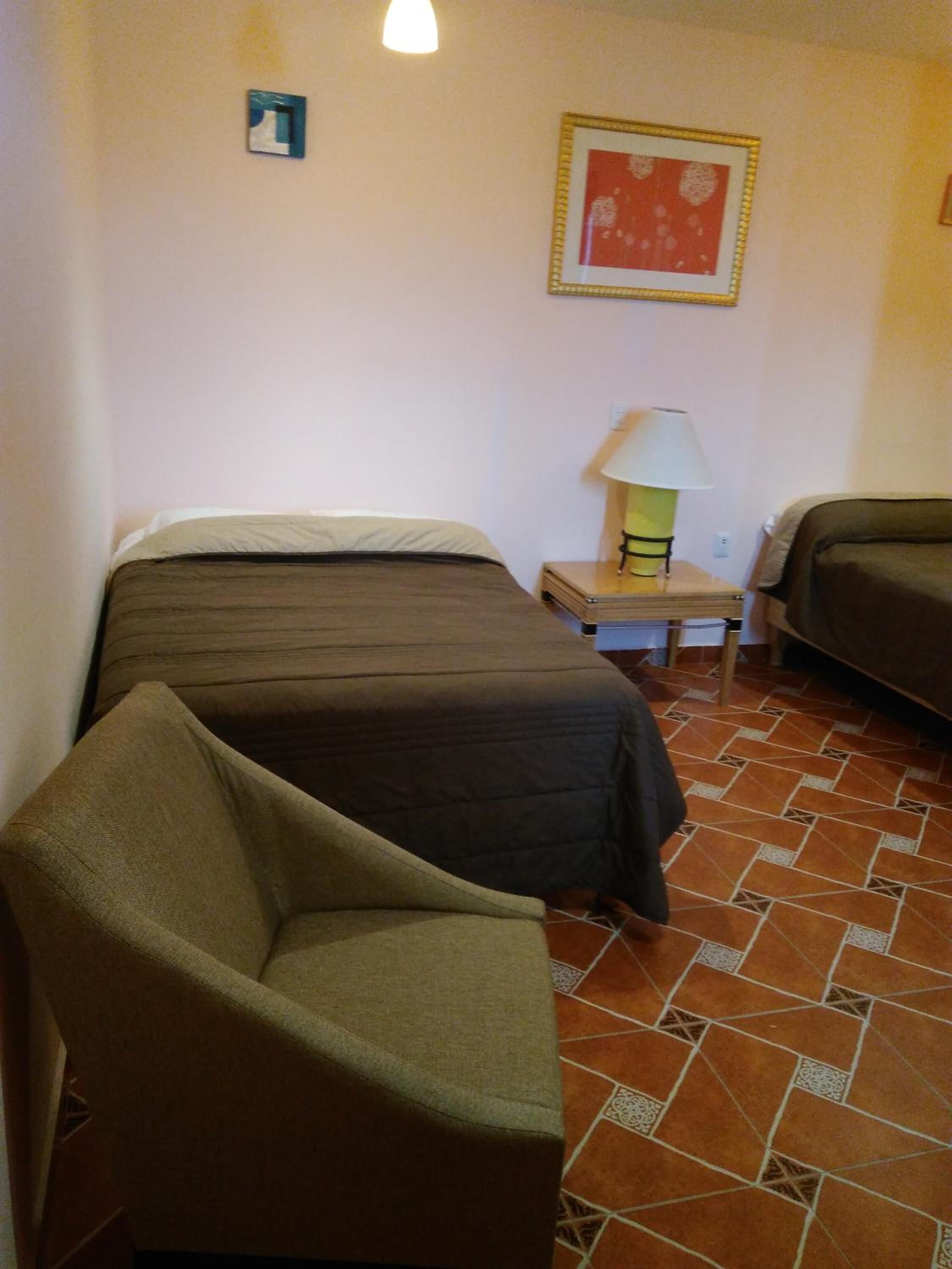 A room with two beds, a chair, a nightstand with a lamp, and a patterned floor.