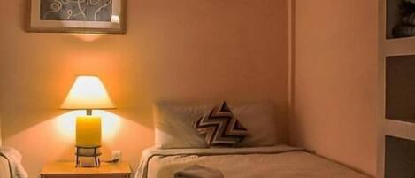 A hotel room with a bed, a bedside table with a lamp, a framed artwork on the wall, and a small shelf with items.