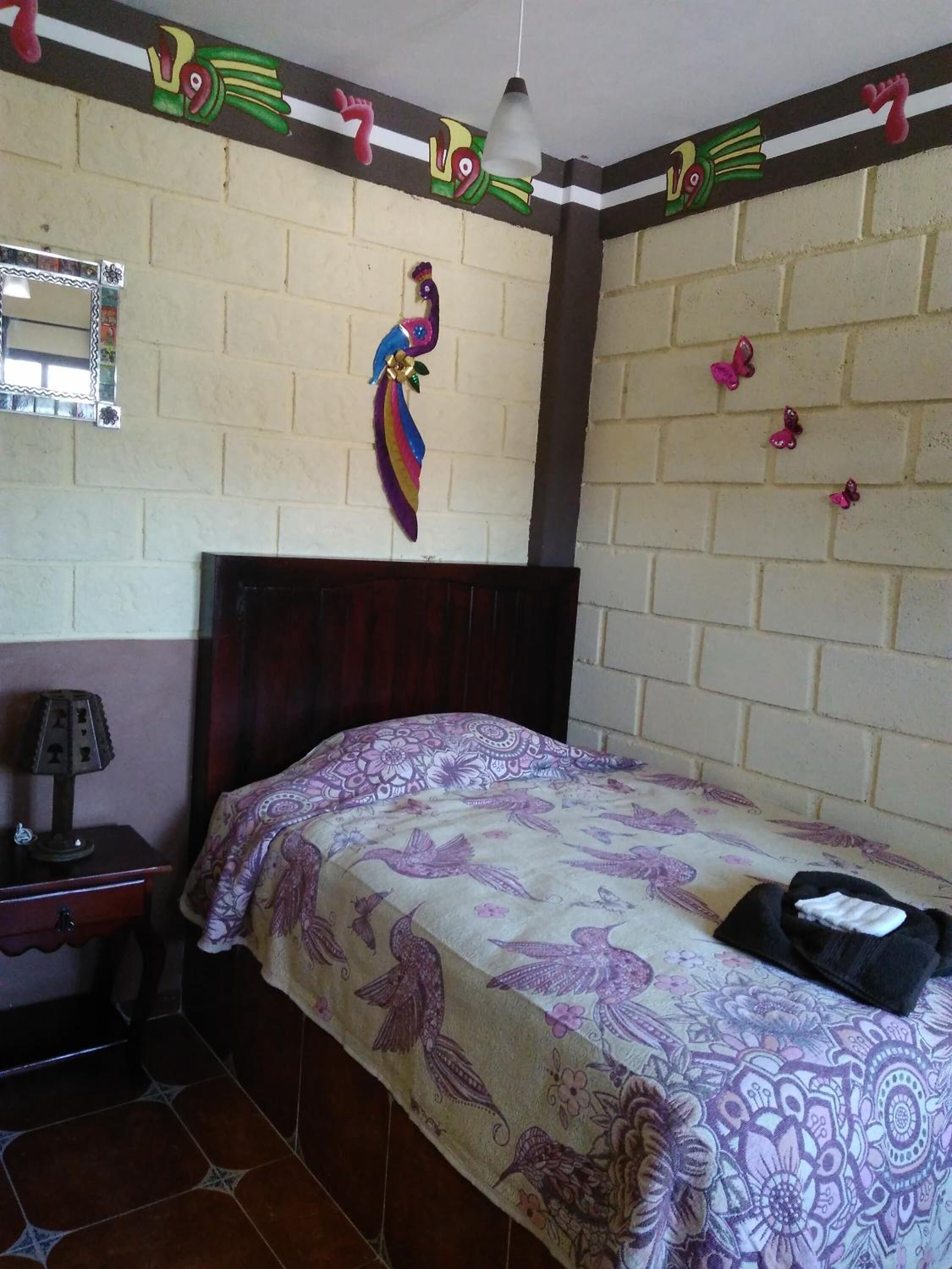 A room with a bed, a nightstand, a lamp, a mirror, and a wall with colorful decorations.