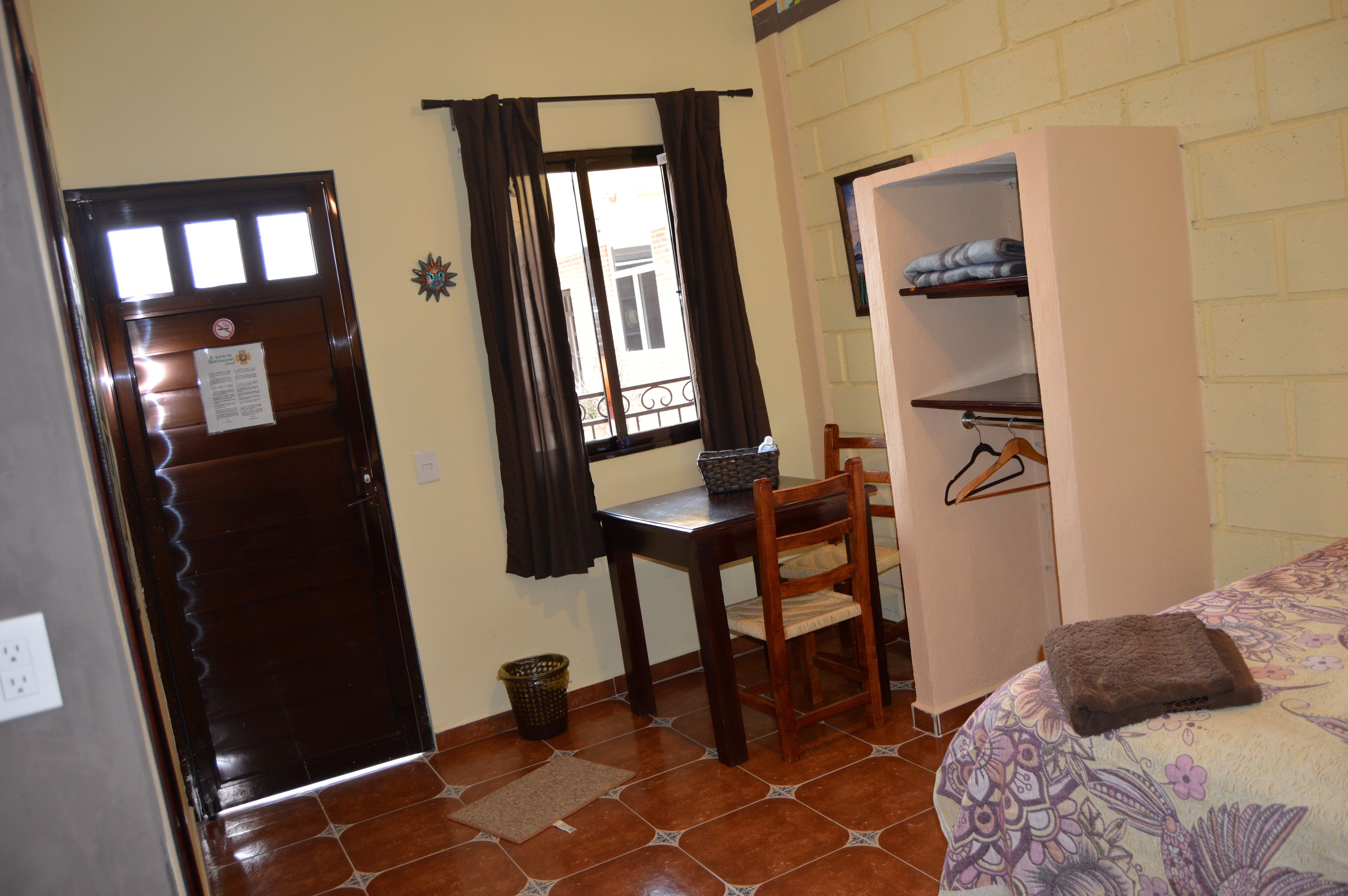 Une pièce avec une porte en bois, une petite table et une chaise, une armoire et un lit recouvert d’une courtepointe à motifs floraux.