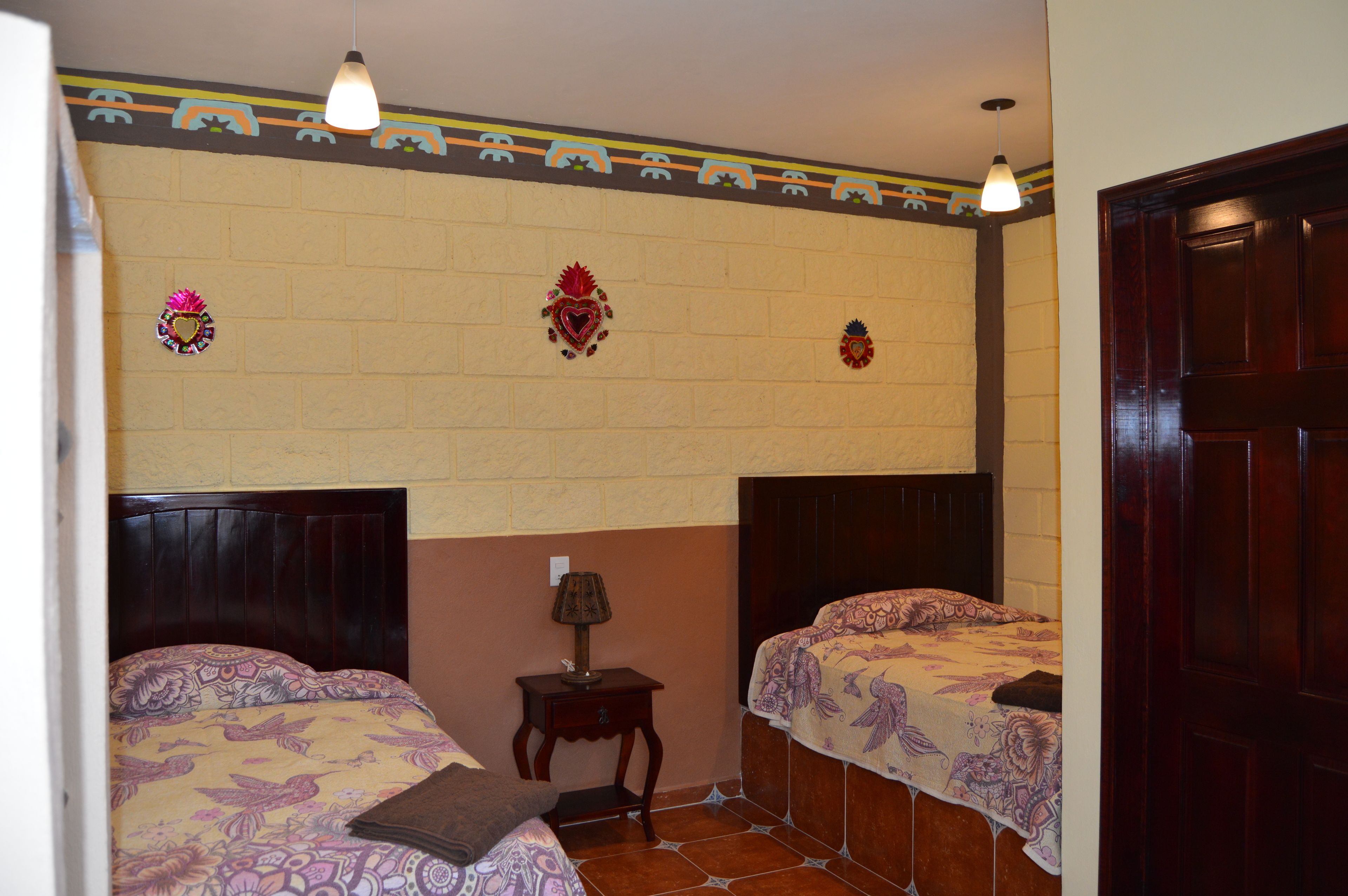 Een kamer met twee bedden, een klein tafeltje met een lamp, decoratieve wandversieringen en een houten deur.