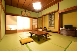 Una habitación japonesa tradicional con suelo de tatami, una mesa de madera baja, puertas correderas y un televisor sobre un mueble de madera.