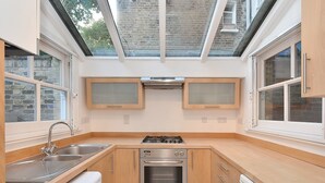 A compact kitchen with a gas stove, oven, and built-in washing machine. It features wooden cabinets, a window, and a skylight. - Belgravia Apartments - Cathcart Road (London)
