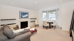 A living room with a fireplace, a sofa, a dining table with chairs, and a large window with decorative valance. - Belgravia Apartments - Cathcart Road (London)