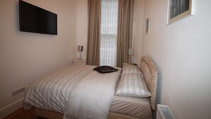 A bedroom with a bed, a flat-screen TV mounted on the wall, a window with curtains, and a radiator. - Belgravia Apartments - Gloucester Road (London)