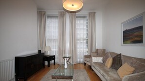 A living room with a sofa, a coffee table, a side table, a lamp, and a painting on the wall. - Belgravia Apartments - Gloucester Road (London)