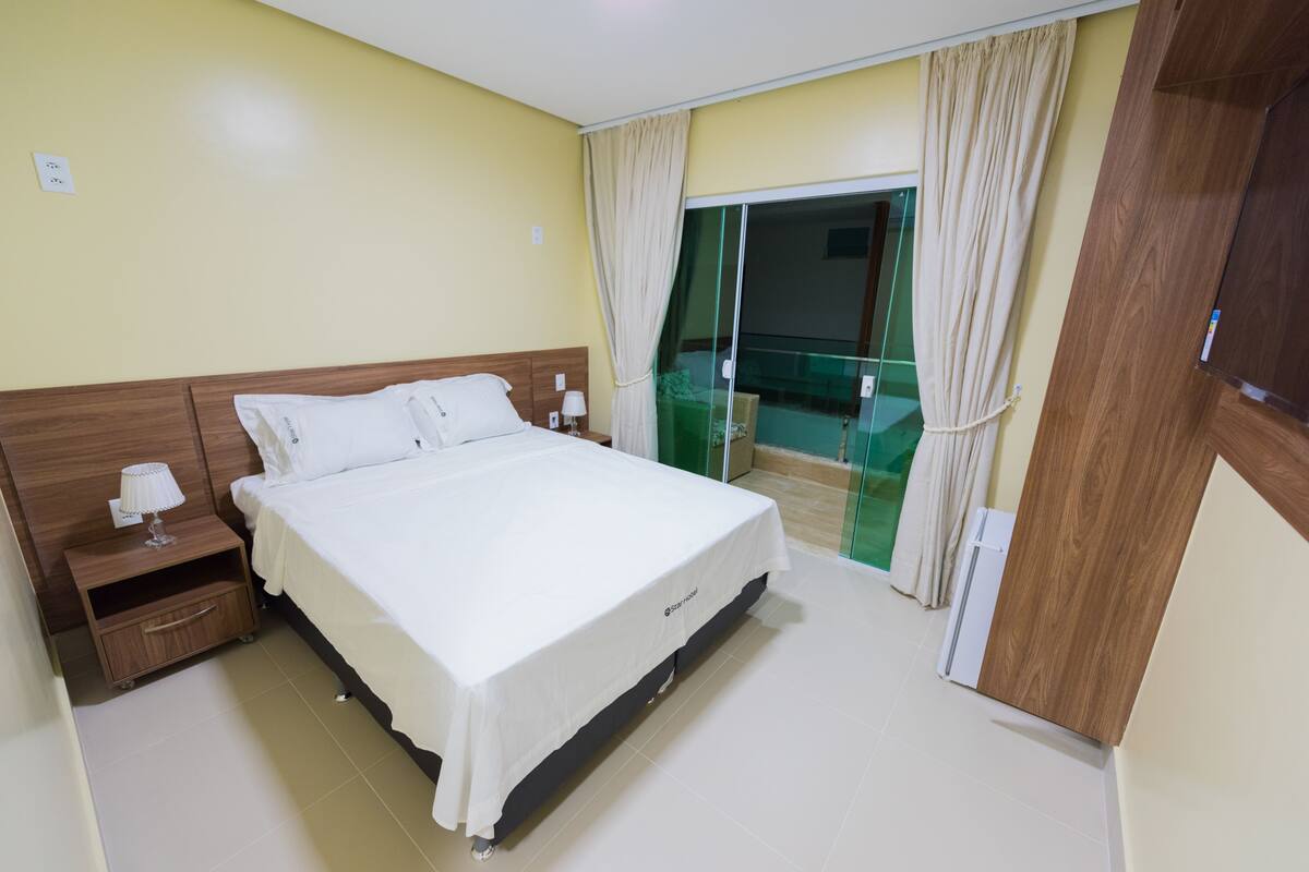 A hotel room with a large bed, wooden headboard, bedside tables with lamps, and a sliding glass door leading to a balcony.