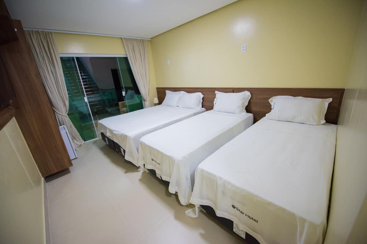 A hotel room with three single beds, a sliding glass door leading to a balcony, and a wooden wardrobe.