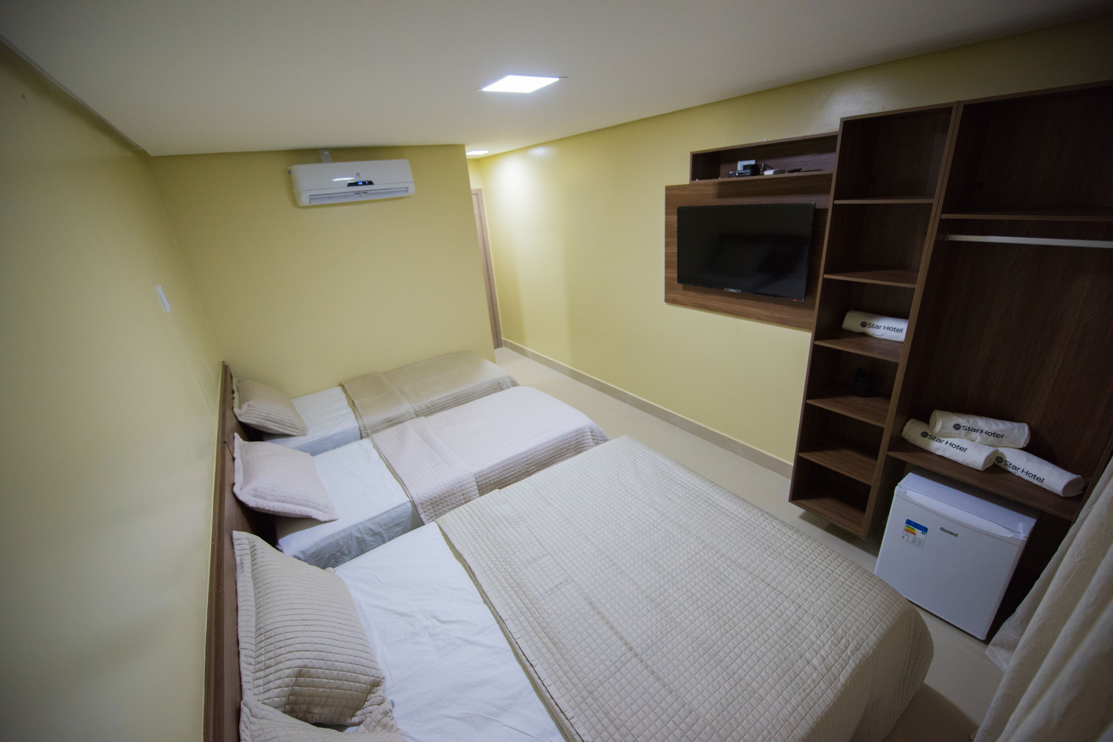 A hotel room with two beds, a TV mounted on the wall, a wardrobe, and a small refrigerator.