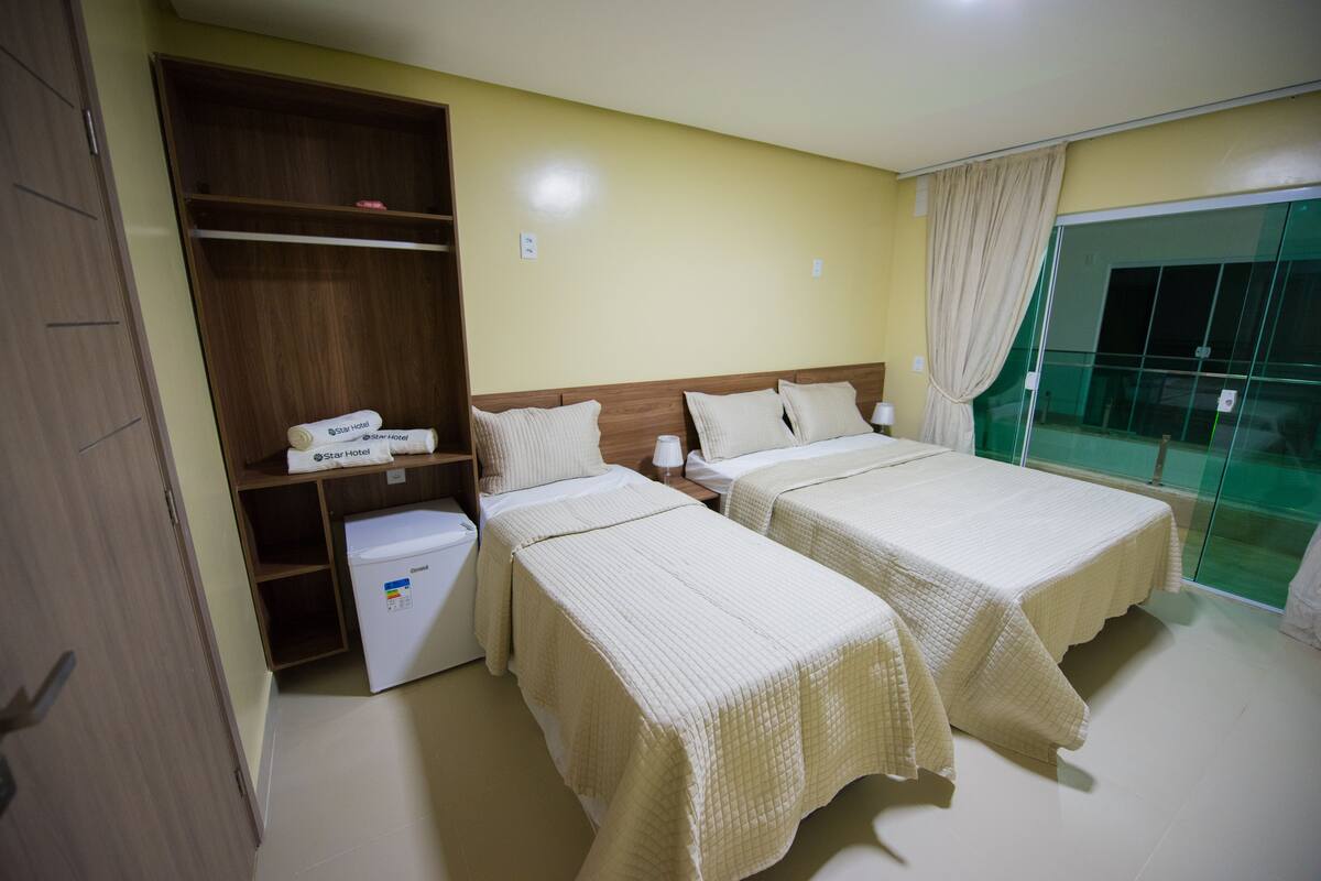 A hotel room with two beds, a wardrobe, a mini-fridge, and a view of the outside.