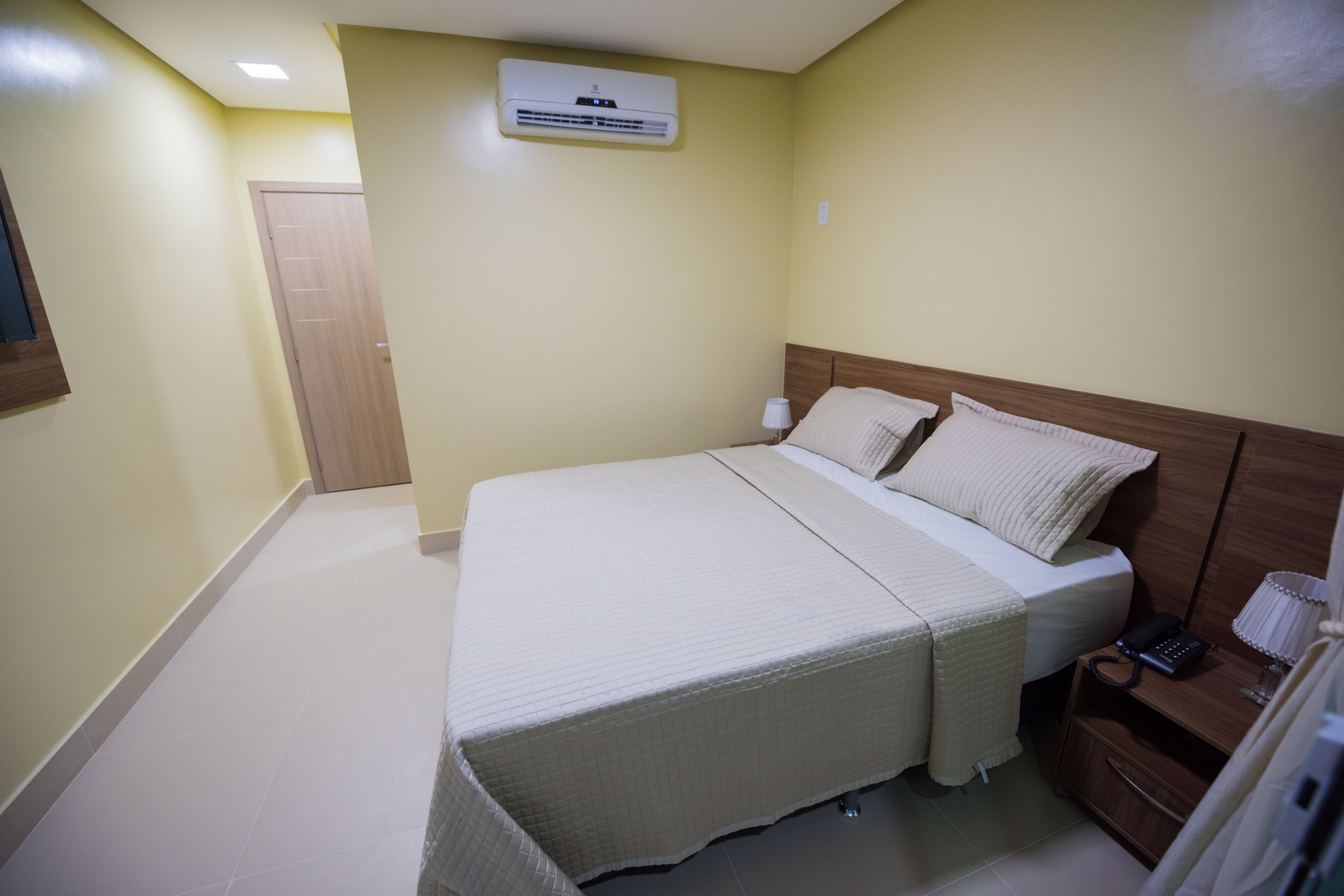 A bedroom with a bed, bedside table, lamp, and an air conditioning unit.