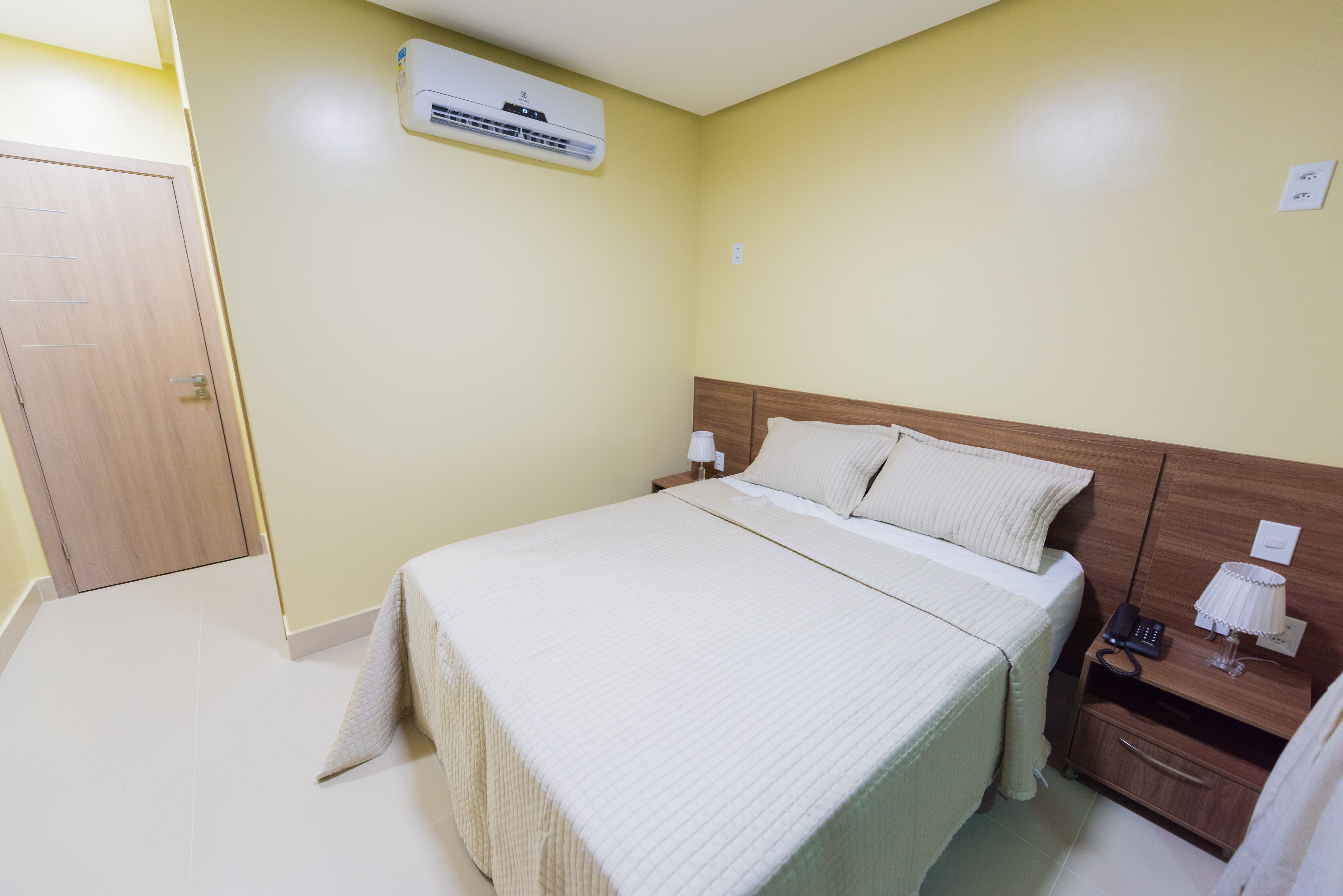 Een hotelkamer met een bed, nachtkastje, lamp en een airconditioner aan de muur.