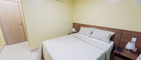 A hotel room with a bed, bedside table, lamp, and an air conditioning unit on the wall.