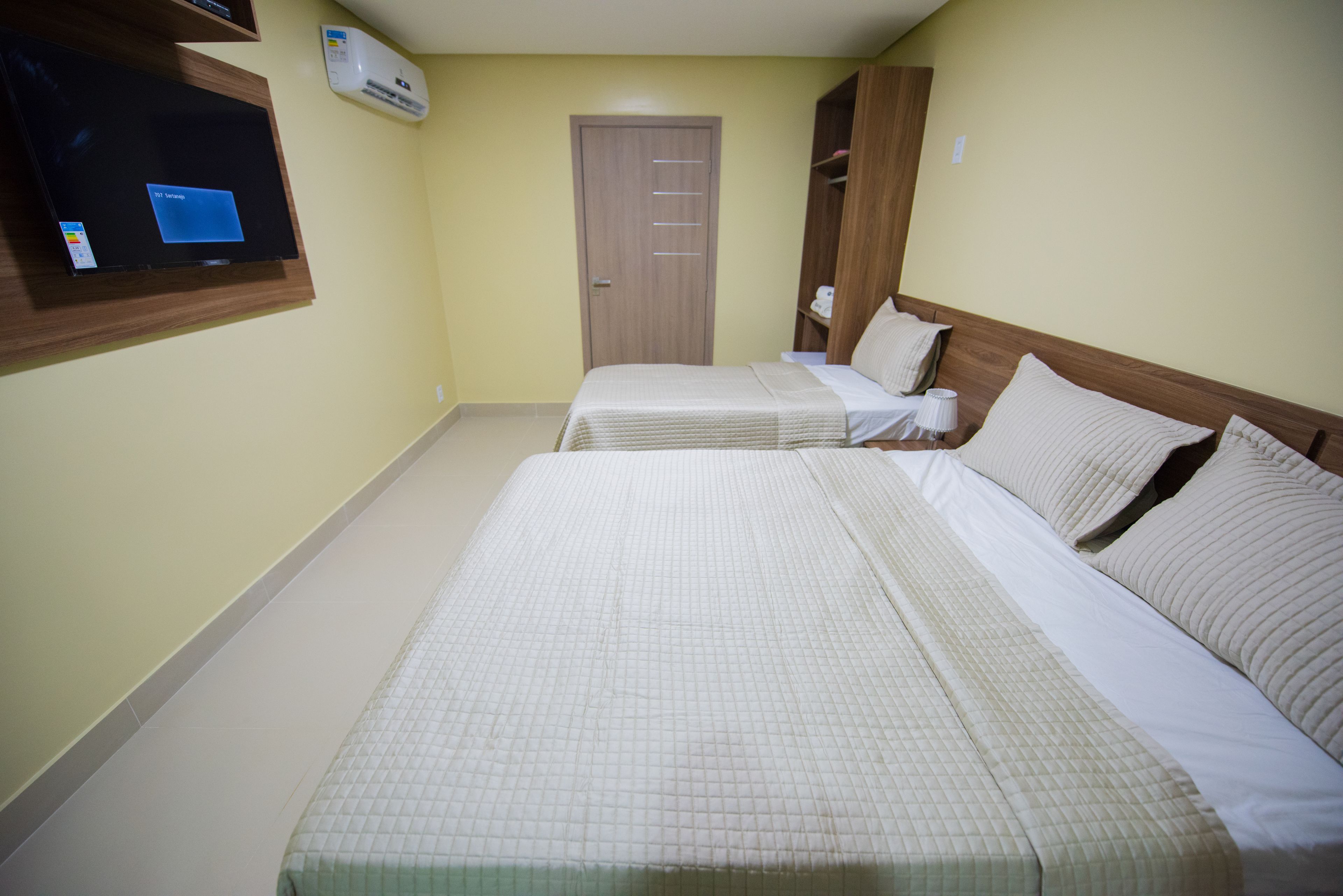 A hotel room with two beds, a TV mounted on the wall, an air conditioning unit, and a door.