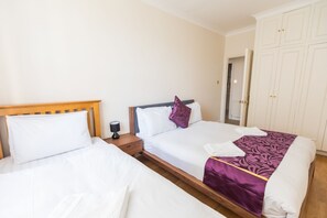 Quarto de hotel com duas camas de solteiro, cabeceiras de madeira, roupa de cama branca e um cobertor roxo, além de um guarda-roupa com puxadores dourados.