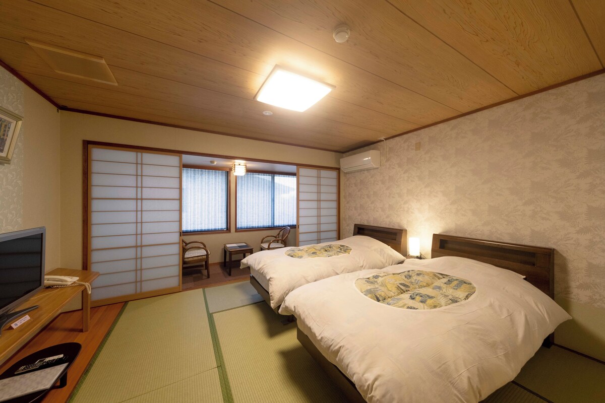 A traditional Japanese-style bedroom with tatami flooring, two beds with floral bedding, a desk with a telephone, and sliding shoji doors.