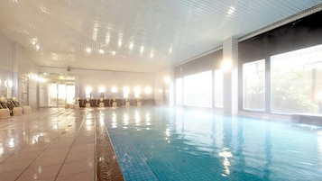 Una piscina interior con paredes y suelo de baldosas, zona de vestuarios con bancos, y grandes ventanales que permiten la entrada de luz natural.